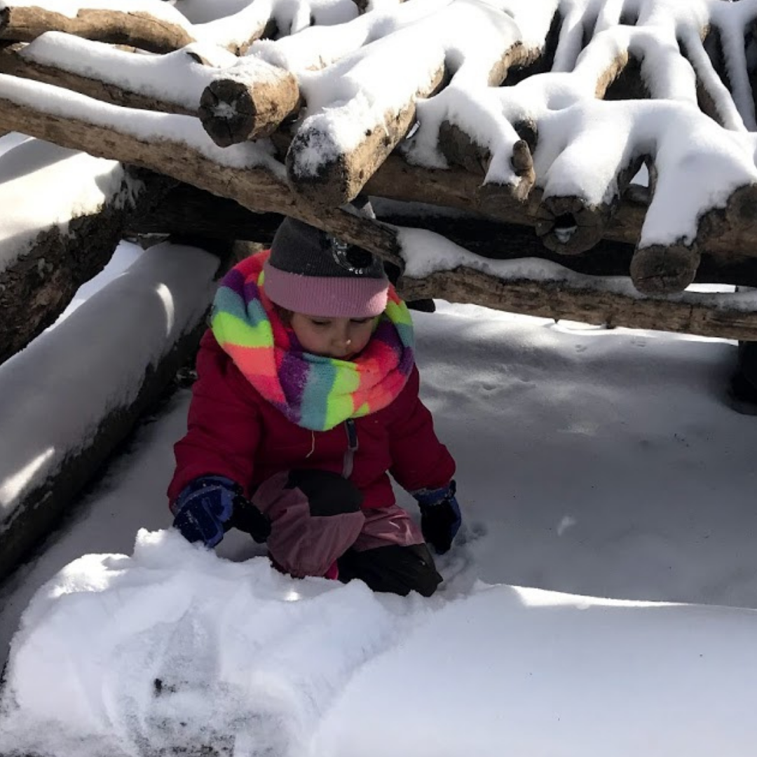 A young child plays in the snow in a fort made from logs and branches. They are wearing a bright, multicolor scarf, a grey sock hat with a pink brim, and a dark pink coat, They are surrounded by snow. 